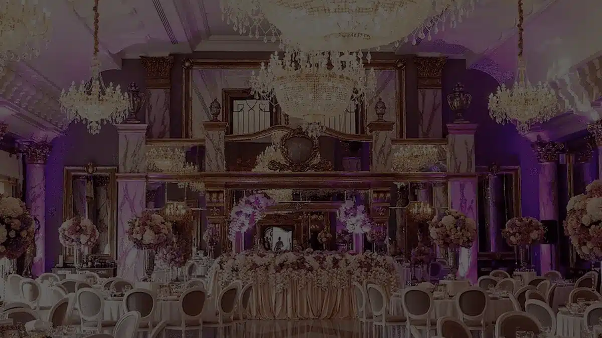 Ballroom with crystal chandeliers, purple uplit pillars and pink flower centerpieces on white tables