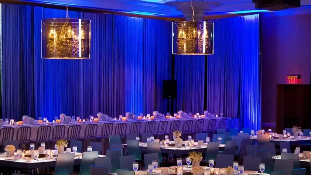 Reception room with blue-lit drapes, glowing chandeliers and long tables set for a party