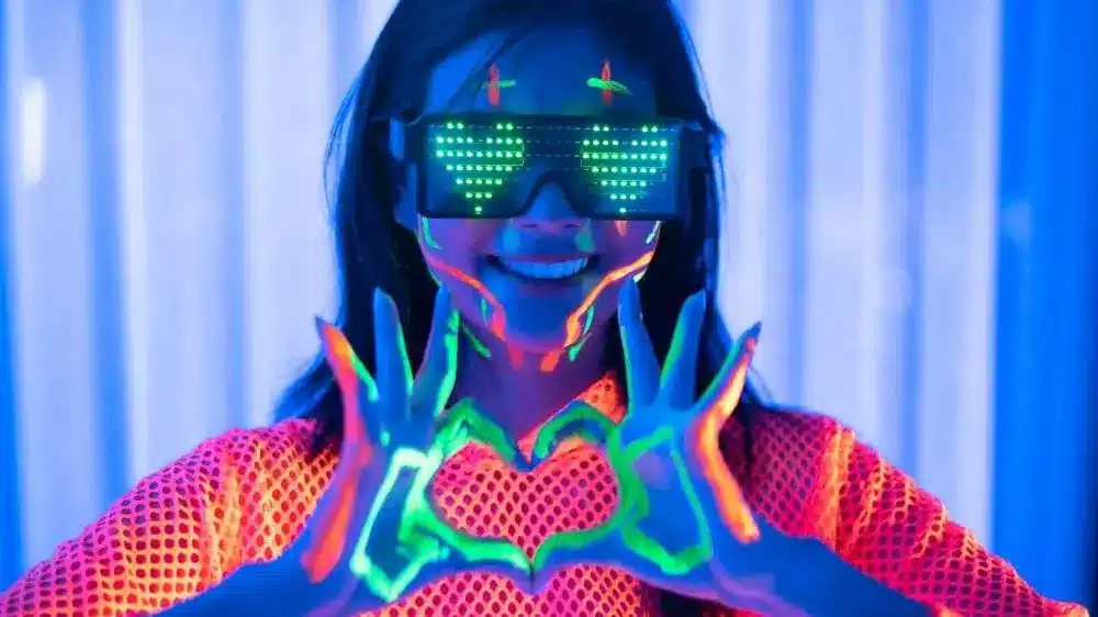 Guest in neon face paint and LED glasses making a heart with her hands at a black light party