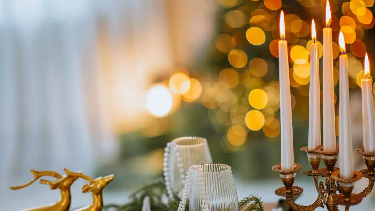 Lit candles in a brass candelabra, gold deer figurines and glasses on a table with Christmas tree lights behind