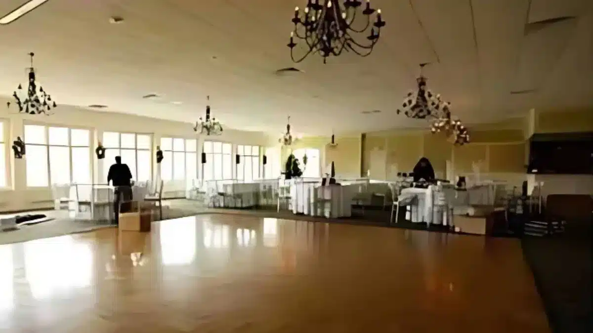 Plain banquet hall with chandeliers, tall windows and white-covered tables before any event lighting