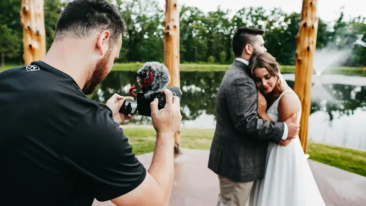 Camera operator filming a bride and groom embracing under a wooden arbor at an outdoor wedding