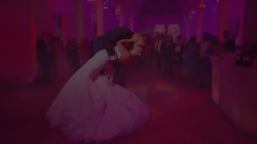 Couple dancing their first dance in a reception hall washed in pink uplighting, guests watching in the background
