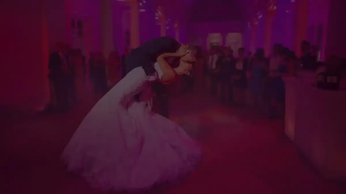 Couple dancing their first dance in a reception hall washed in pink uplighting, guests watching in the background