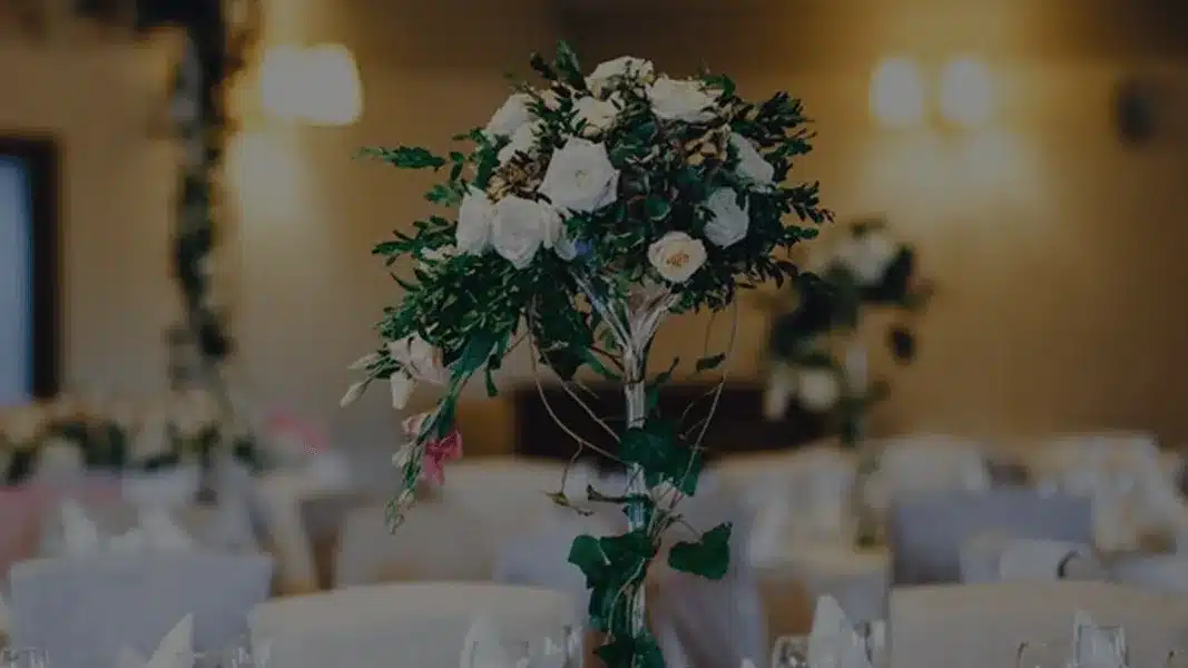 Tall arrangement of white roses and greenery above white-covered reception tables in a softly lit hall
