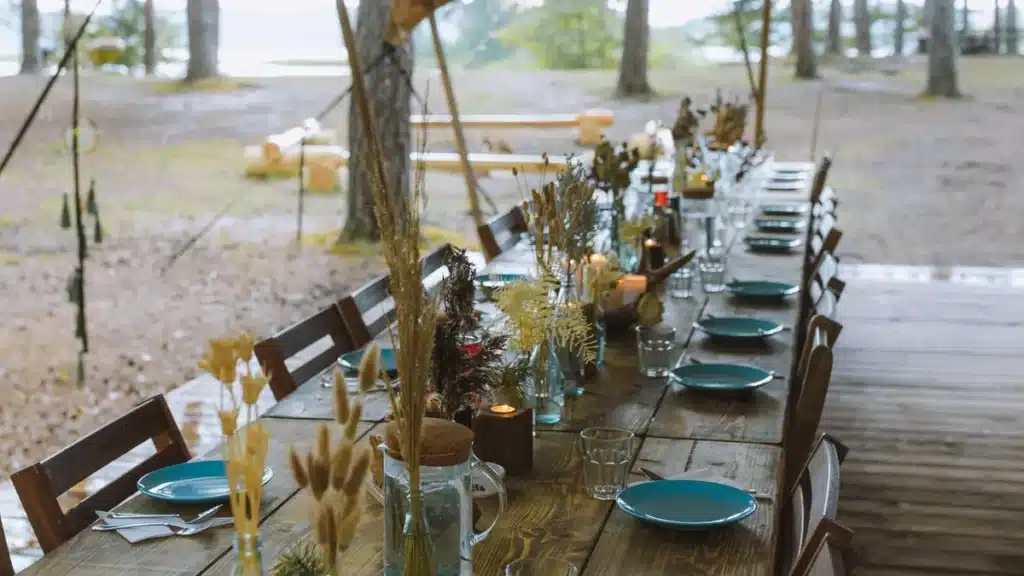 Long wooden table set with teal plates, candles and dried grasses under a canopy among pine trees