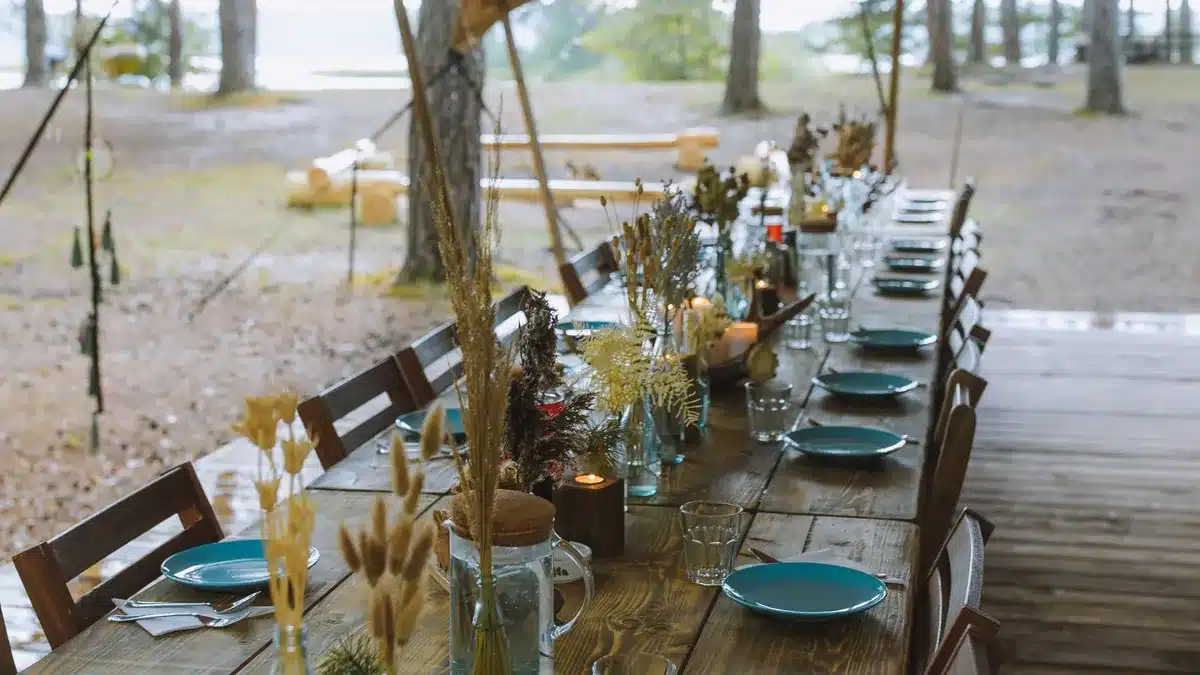 Long wooden table set with teal plates, candles and dried grasses under a canopy among pine trees