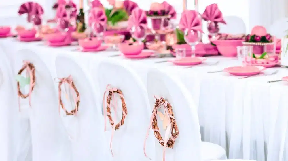 Long table set with pink napkins and plates, with white spandex chair covers decorated with small wreaths