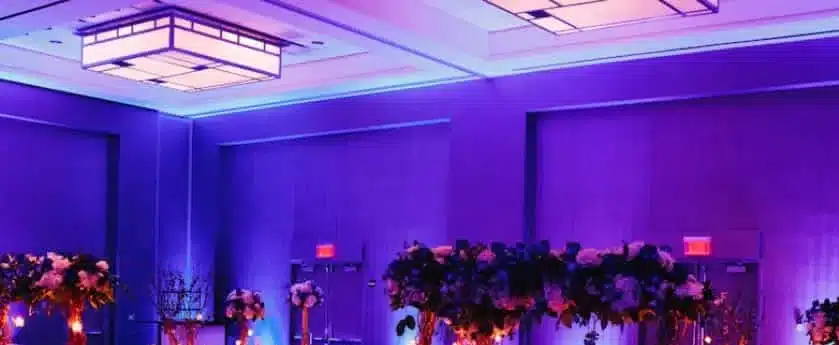 Ballroom walls washed in purple uplighting above tables with tall flower centerpieces