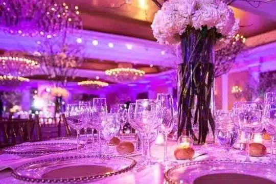 Reception table with tall white flowers, glassware and gold-rimmed chargers under purple light