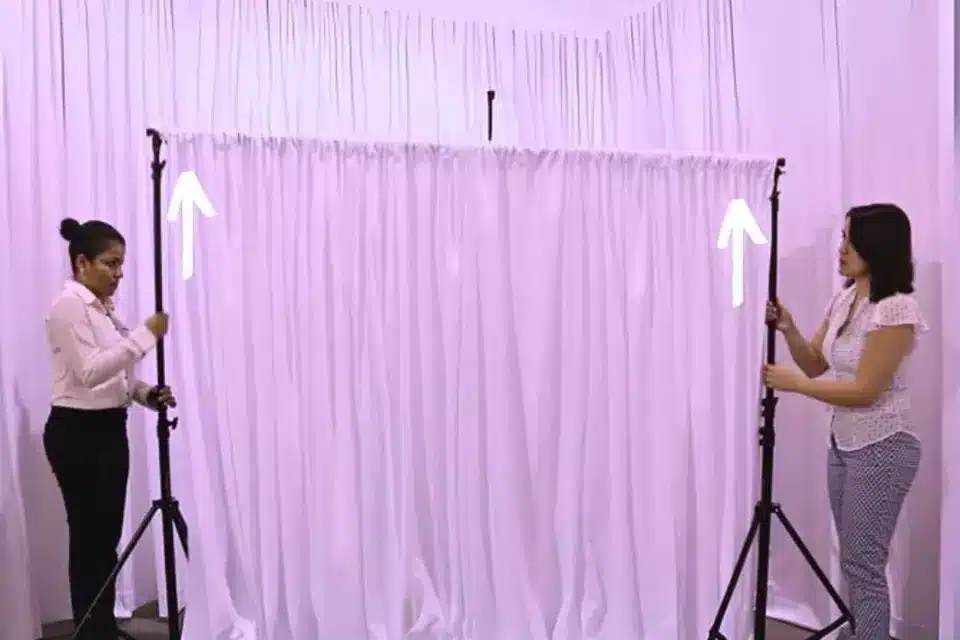 Two people raising the tripod stands of a white pipe and drape backdrop to full height