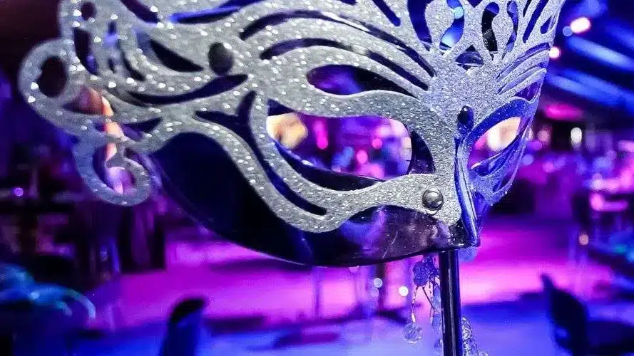 Silver glitter masquerade mask in front of a dim room lit blue and purple