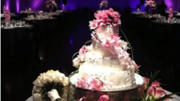 Tiered white wedding cake decorated with pink flowers in a dark reception room with purple uplighting