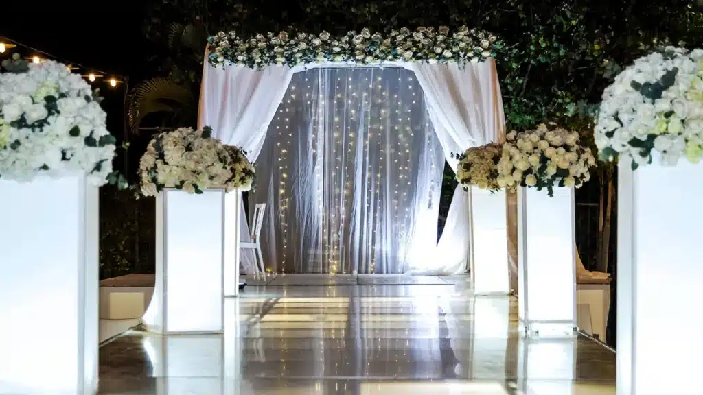 Wedding ceremony canopy with sheer drapes and string lights between white flower columns