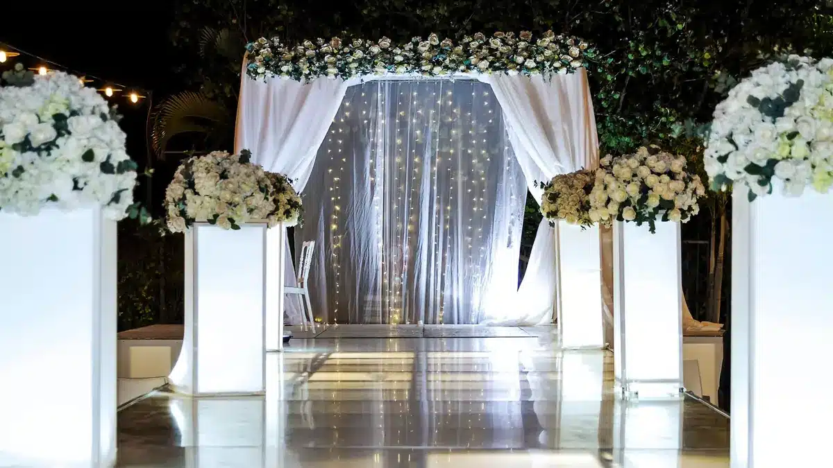 Wedding ceremony canopy with sheer drapes and string lights between white flower columns