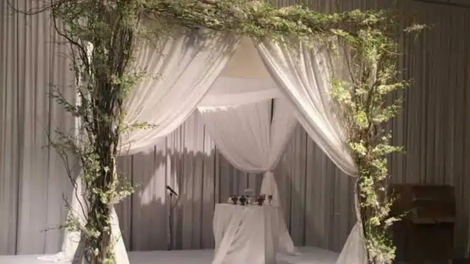 Four-post wedding canopy draped in white fabric and wrapped with trailing greenery