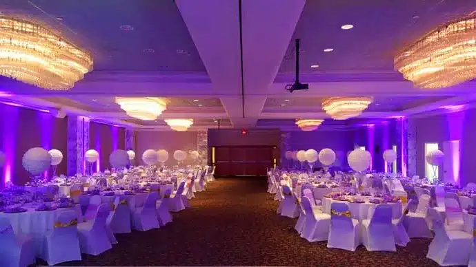 Ballroom with white chair covers and round tables, its walls washed in purple uplighting